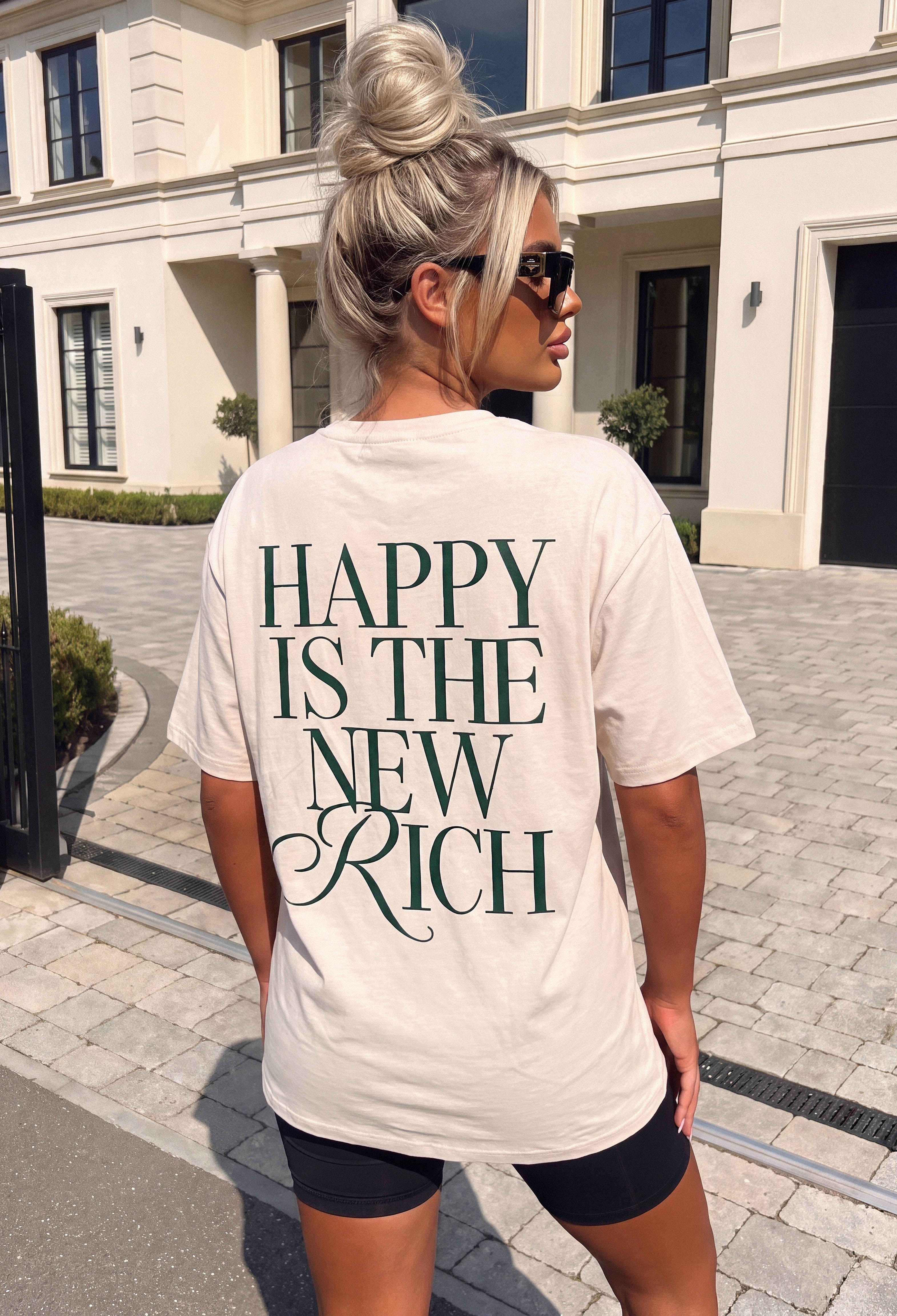Back view of a beige graphic T-shirt with bold green Happy Is The New Rich slogan and relaxed fit.