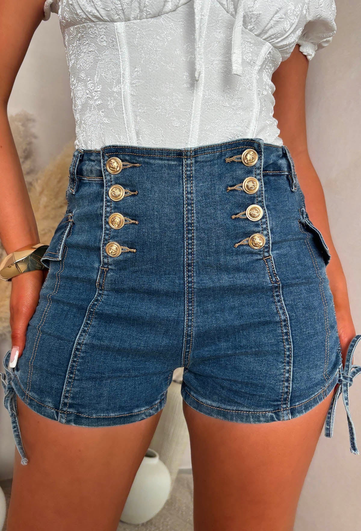 Mid wash stretch denim shorts with a high-waisted fit, gold button front and side tie accents.
