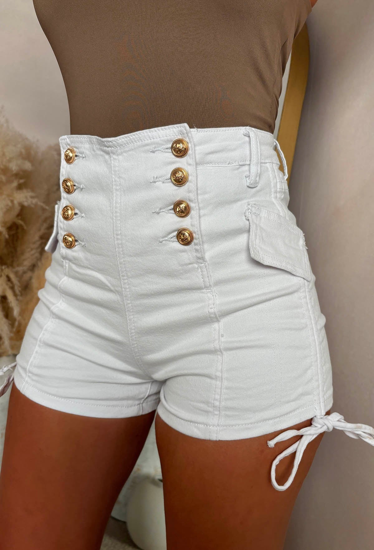 White stretch denim gold button shorts with a high-waisted fit, front gold button detail and adjustable side ties.